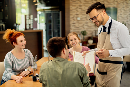 Happy waiter showing menu to a guest who came with friends in a cafe.